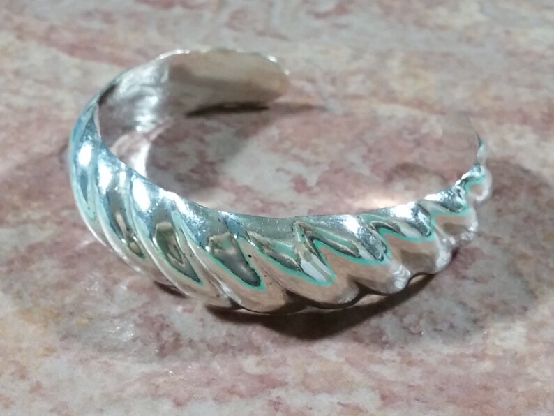 May include: A silver cuff bracelet with a textured, wavy design. The bracelet is open and has a smooth, polished finish.