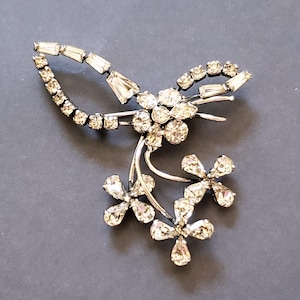 May include: A silver tone brooch with a floral design featuring clear rhinestones. The brooch has a delicate, intricate design with multiple flower shapes.