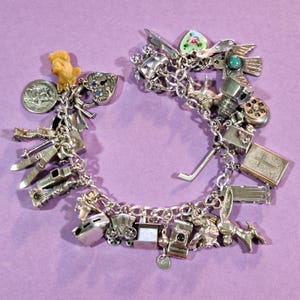 May include: A vintage silver charm bracelet with various charms, including a heart, a cross, a train, a camera, and a bird. The charms are attached to a silver chain. The background is a solid purple color.