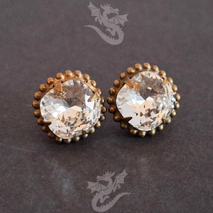May include: A pair of clear, faceted crystal stud earrings with a vintage-style, antique gold-colored setting. Each earring features a large, oval-shaped crystal surrounded by a beaded border. The earrings are displayed on a dark gray background.