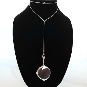 May include: A silver-toned lorgnette necklace featuring a long, delicate chain that connects to a circular, folding magnifying glass pendant. The pendant has an ornate handle and a polished metal frame, suspended from a thin, metallic chain.