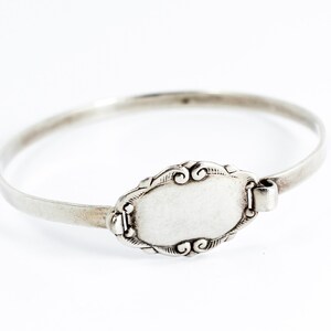 May include: A silver bangle bracelet with a blank oval charm. The charm has a decorative border.