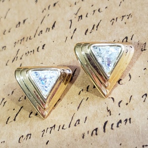 May include: A pair of gold-toned triangle stud earrings. Each earring features a clear, triangular-cut stone set within a gold frame. The earrings have a layered design and a polished finish, reflecting light.