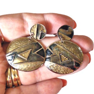 May include: Gold-toned, geometric earrings with black and gold accents. The earrings have a circular design with a smaller circle at the top. The earrings are held in a hand.