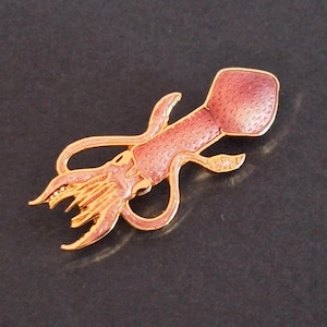 Könnte beinhalten: Goldfarbene Tintenfisch-Brosche mit rosa Emaille-Details. Die Brosche zeigt ein detailliertes Design eines Tintenfischs mit Tentakeln und einem strukturierten Körper. Der Tintenfisch ist vor einem dunklen Hintergrund platziert.
