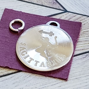 Op de afbeelding: Zilveren bedel met een Boogschutter-sterrenbeeld en het woord "Boogschutter" gegraveerd rond de rand. De bedel heeft een springring om aan een ketting of armband te bevestigen.