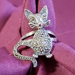 May include: A silver ring with a detailed cat design. The cat is covered in small, sparkling stones. The ring is on a purple fabric background.
