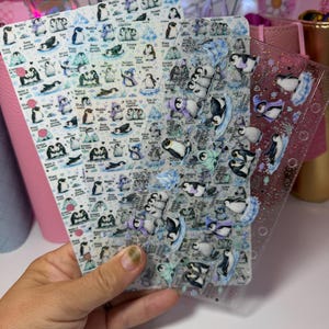 May include: Three clear plastic planner dividers featuring a repeating pattern of illustrated penguins in various poses, some wearing scarves or hats, interspersed with small blue snowflakes and inspirational text phrases like Dream big and Stay cool.