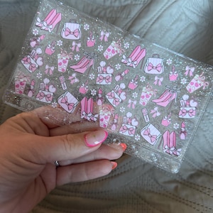 May include: Clear, rectangular planner cover with pink and white illustrations of high heels, purses, bows, and lipstick. The cover is decorated with glitter and has a feminine theme.