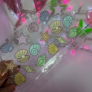 May include: A clear, rectangular plastic binder cover featuring a repeating pattern of pastel-colored seashells and starfish. The transparent material is embedded with fine silver glitter and small star-shaped confetti, with six circular holes punched along one side.