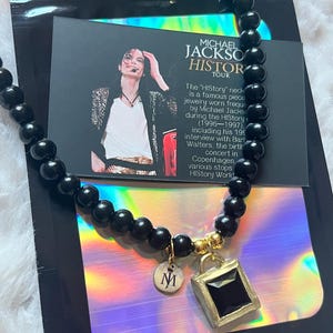 Puede incluir: Un collar con cuentas negras lisas y un colgante dorado con una piedra negra cuadrada. Un pequeño dije redondo dorado con la letra M está unido junto al colgante. El collar se muestra sobre una tarjeta con la imagen de Michael Jackson.