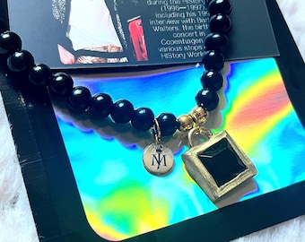 May include: A necklace featuring black spherical beads, a gold-toned metal charm with the letter M, and a gold-toned square pendant with a black center stone. The necklace is displayed on a card with text about the Michael Jackson HIStory tour.
