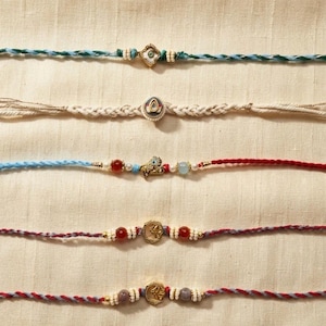 May include: Five braided cord bracelets featuring central gold-tone metal charms, small beads, and colorful threads. The designs include a diamond-shaped charm, a teardrop shape, a bird figure, and two circular medallions with red and grey beads.