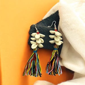 Peut inclure: Boucles d'oreilles pendantes avec des perles blanches en forme de pétales et des pompons colorés. Les boucles d'oreilles ont des crochets argentés et sont présentées sur un fond sombre, avec un tissu blanc et un fond orange.