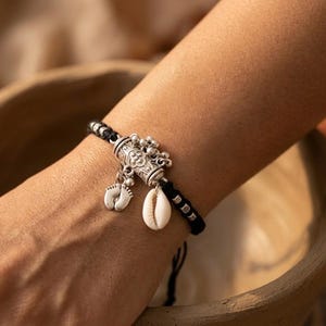 Può includere: Un bracciale in corda nera con un ciondolo cilindrico color argento con incisioni floreali, piccoli campanellini argentati pendenti, un ciondolo color argento a forma di piedini di neonato e una conchiglia bianca fissata al cordino.