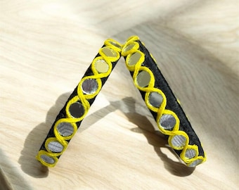 May include: Two black bangles with yellow trim and small, round, mirrored accents. The bangles are arranged diagonally on a light wood surface, with one slightly overlapping the other. The mirrored accents reflect light, adding a touch of sparkle.