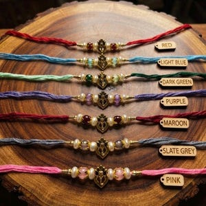 May include: Seven bracelets with colored threads in red, light blue, dark green, purple, maroon, slate grey, and pink. Each features a central gold-toned Ganesha charm flanked by white beads and colored stones, with wooden labels identifying each color.