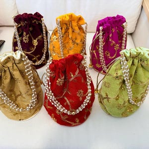 Può includere: Una collezione di sette borse a coulisse in velluto di vari colori, tra cui oro, rosso, giallo, viola e verde. Ogni borsa è adornata da ricami dorati e un cinturino di perline. Le borse sono disposte su una superficie bianca.
