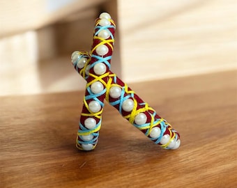 May include: Two decorative, handcrafted sticks in a cross shape. The sticks are wrapped in red fabric and adorned with yellow, light blue, and white thread, along with white pearl beads. The sticks are resting on a wooden surface.