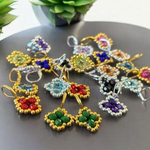 May include: Assortment of colorful beaded earrings with various gemstone colors, including green, red, blue, purple, and black. Each earring features a square-shaped design with a dangling drop.
