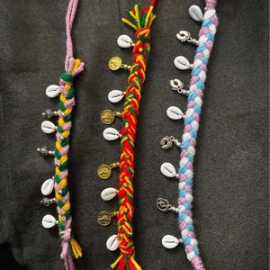 May include: Three braided anklets in the image. One is pink, yellow, and green with silver charms and white shells. The second is red, yellow, and green with gold coins and white shells. The third is blue, white, and pink with silver charms and white shells.