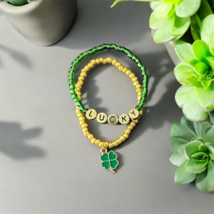 Könnte beinhalten: Ein goldfarbenes und grünes Perlenarmband mit einem Glücksbringer aus vierblättrigem Kleeblatt und dem Wort "LUCKY" in goldenen Perlen.