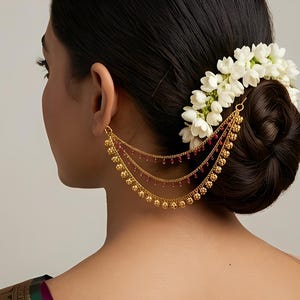 Puede incluir: Pendiente de oro con tres cadenas en cascada, adornado con pequeñas cuentas rojas y esferas doradas decorativas. La joya está sujeta a la oreja y cae hacia abajo. El cabello del modelo está recogido en un moño y decorado con flores blancas.