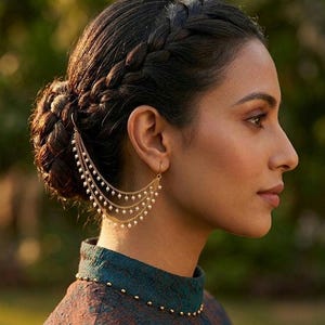 May include: A gold-toned ear cuff with three cascading strands of small, white pearls. The jewelry is attached to the ear and drapes down. The person is wearing a teal and brown patterned top.