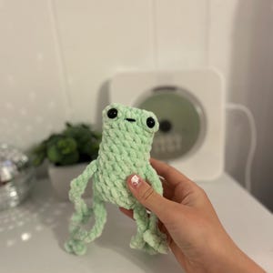 May include: A light green crocheted frog plushie with black eyes and a small mouth. The frog has long, dangling arms and legs. The plushie is made of a soft, textured yarn. The background includes a white surface and a plant in a white pot.