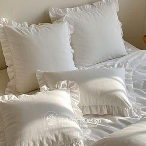 May include: A set of four white ruffled pillowcases. The pillows are arranged on a white bedspread. The pillows are all made of a soft, white fabric and have a delicate ruffle trim.