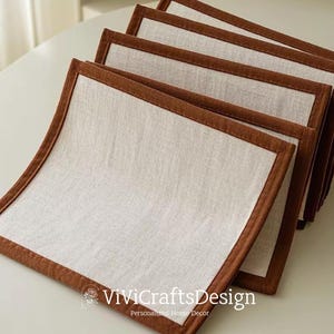 Op de afbeelding: Set van zes bruine en witte placemats met een bruine suède rand. De placemats zijn gemaakt van natuurlijke linnen stof.