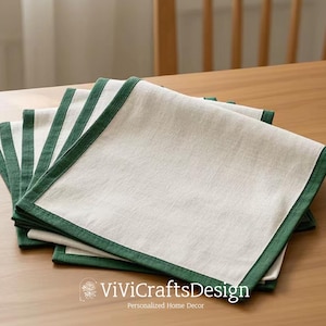 Könnte beinhalten: Ein Stapel weißer Leinenservietten mit grünem Rand. Die Servietten sind in der Hälfte gefaltet und übereinander gestapelt. Die Servietten liegen auf einem Holztisch.