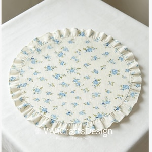 Op de afbeelding: Ronde, crèmekleurige placemat met een blauw bloemenpatroon en een gerimpelde rand. De placemat heeft kleine blauwe bloemen verspreid over het oppervlak. De tekst "ViviCraftsDesign" staat onderaan. Een decoratief item voor in huis.