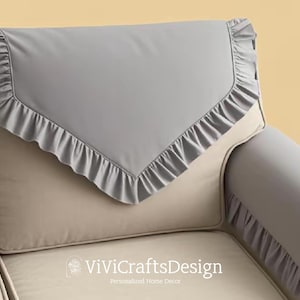 May include: A gray couch with a gray and white ruffled throw pillow and matching armrest covers. The couch is made of a soft, plush fabric and has a classic design.