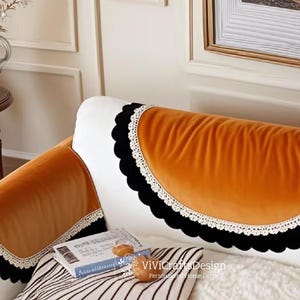 May include: Orange velvet sofa arm covers with black scalloped edges and white lace trim. The covers are placed on a white sofa with a striped pillow and a book. The overall theme is home decor.