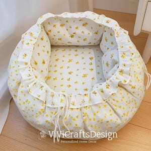 May include: A round pet bed with a white base and a yellow floral pattern. The bed has a ruffled edge and a drawstring closure. The interior is also patterned with yellow flowers. The bed is designed for comfort and style.