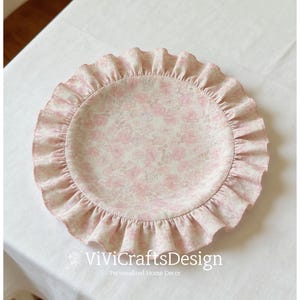 Può includere: Una tovaglietta rotonda in tessuto con un motivo floreale rosa tenue e bianco e un bordo arricciato a volant. Il centro presenta una fitta stampa di rose, mentre il bordo è costituito da una balza in tessuto plissettato dello stesso materiale.