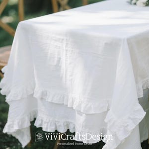 May include: A white linen tablecloth with ruffled edges. The tablecloth is draped over a table, showcasing its texture and design. The image is well-lit, highlighting the fabric's details. The tablecloth is suitable for home decor.