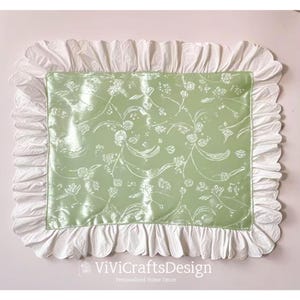 May include: A light green pillow with a white floral pattern and a white ruffled border. The pillow is made of satin and has a soft, luxurious feel.