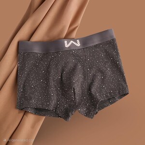 May include: Dark grey boxer shorts with a white speckled pattern. The waistband is a darker grey with a white logo. The shorts are made of a soft material and are designed for comfort.