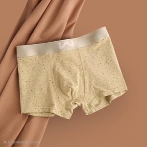 May include: Light yellow boxer shorts with a speckled pattern and a beige waistband. The waistband features a white logo. The shorts are made of a soft material and are displayed on a brown background.