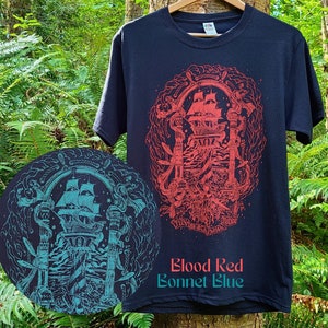 A black tshirt with a detailed illustration of pirate iconography. Illustrated typography at the bottom of the design reads: fun with friends at sea. The design is screen printed with red ink. A circle in the corner shows an alternative blue ink.