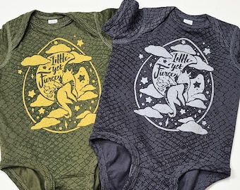 Puede incluir: Dos bodies de bebé, uno verde y otro negro, con un estampado gráfico de un dragón en una luna con el texto "Little yet Fierce".
