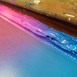 Puede incluir: Primer plano de un proceso de serigraf&iacute;a con colores vibrantes. Un marco de madera sostiene una pantalla con un degradado de tinta rosa, morado y azul. La tinta es gruesa y texturizada, creando un dise&ntilde;o abstracto visualmente atractivo.