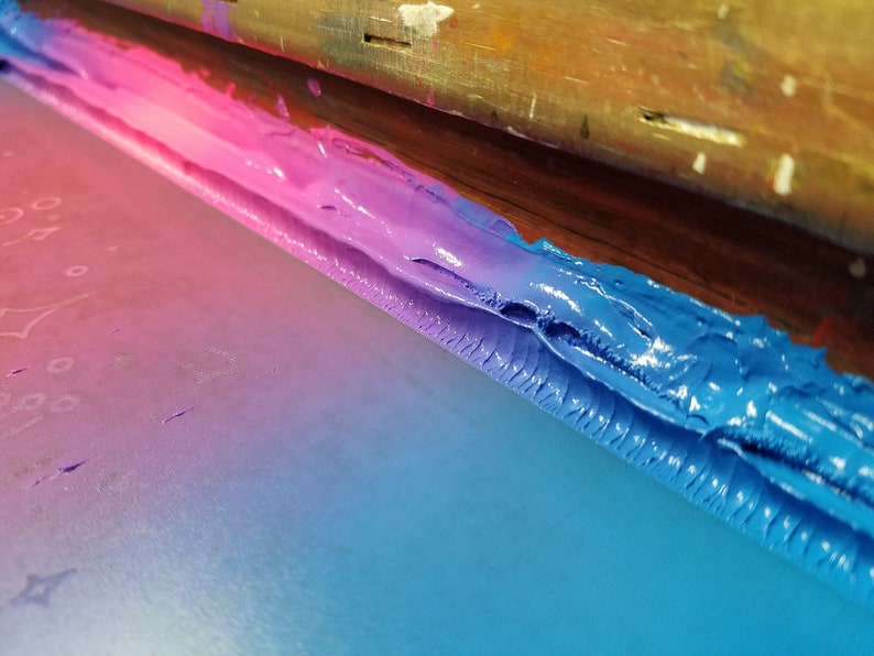 Puede incluir: Primer plano de un proceso de serigraf&iacute;a con colores vibrantes. Un marco de madera sostiene una pantalla con un degradado de tinta rosa, morado y azul. La tinta es gruesa y texturizada, creando un dise&ntilde;o abstracto visualmente atractivo.