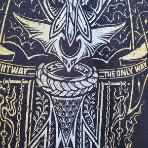 Peut inclure: T-shirt noir avec un motif graphique or et blanc. Le motif repr&eacute;sente une cr&eacute;ature ail&eacute;e avec un cercle de corde et le texte "The Right Way Is Not The Only Way".