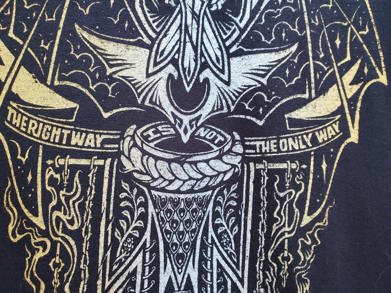 Peut inclure: T-shirt noir avec un motif graphique or et blanc. Le motif repr&eacute;sente une cr&eacute;ature ail&eacute;e avec un cercle de corde et le texte "The Right Way Is Not The Only Way".