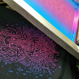 Puede incluir: Una camiseta negra con un dise&ntilde;o gr&aacute;fico en rosa ne&oacute;n y azul es parcialmente visible debajo de un marco de serigraf&iacute;a. El dise&ntilde;o presenta formas geom&eacute;tricas, l&iacute;neas y un emblema central. El marco de serigraf&iacute;a tiene un degradado de azul y rosa.