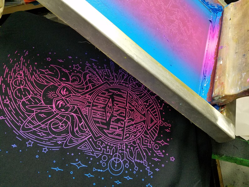 Puede incluir: Una camiseta negra con un dise&ntilde;o gr&aacute;fico en rosa ne&oacute;n y azul es parcialmente visible debajo de un marco de serigraf&iacute;a. El dise&ntilde;o presenta formas geom&eacute;tricas, l&iacute;neas y un emblema central. El marco de serigraf&iacute;a tiene un degradado de azul y rosa.