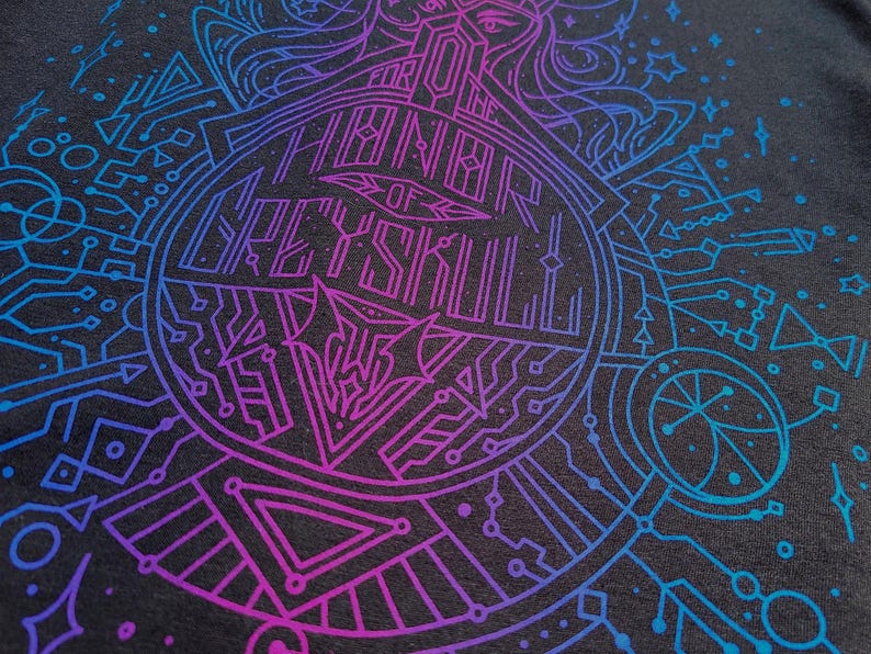 Puede incluir: Camiseta gris oscuro con un dise&ntilde;o geom&eacute;trico vibrante en azul y morado. El dise&ntilde;o presenta un emblema circular con el texto "FOR THE THRONE OF GREYSKULL" y un intrincado arte lineal.